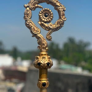 May include: A gold-coloured pastoral staff featuring an ornate, scrolled crook head with a central flower motif. The staff includes a decorative collar adorned with black circular inlays and intricate leaf-like carvings, mounted on a smooth pole.
