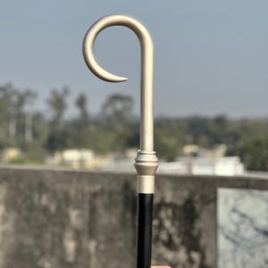Pode incluir: Um cajado de pastor com um gancho de tom prateado fosco e liso e uma peça de colarinho decorativa fixada a uma haste cilíndrica longa, reta e preta sólida. O design é simples e funcional, com um topo curvo e um acabamento liso.