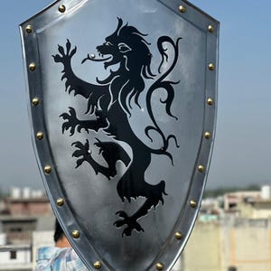 May include: A heater-style shield made of polished silver-toned metal featuring a black rampant lion silhouette in the center. The shield is bordered by a raised rim with evenly spaced gold-toned rivets along the entire outer edge of the frame.