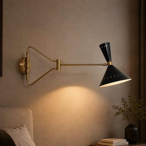 Pode incluir: Uma luminária de parede com braço articulado em latão e cúpula de cone duplo em preto fosco. A cúpula possui pequenas perfurações ao longo da borda e a lâmpada emite uma luz quente direcionada para baixo a partir da sua superfície interna branca.