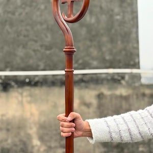 Pode incluir: Um báculo de madeira castanha polida com um bastão longo e reto e uma secção superior curva que forma um laço circular. Dentro do laço circular no topo do bastão, existe uma cruz de madeira esculpida centrada no espaço vazio.