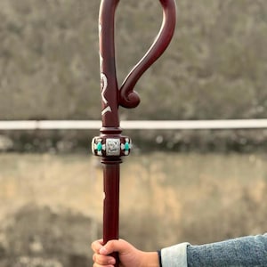 Könnte beinhalten: Ein dunkel rotbrauner Holzstab mit einem gebogenen Haken am oberen Ende und einem dekorativen Kragen. Der Kragen ist mit silberfarbenen Metallakzenten, detaillierten Gravuren und kleinen, ovalen türkis farbenen Einlagen verziert.