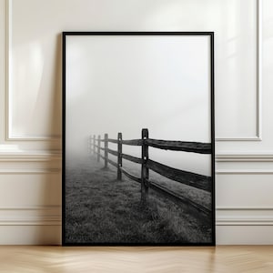 Op de afbeelding: Een zwart-wit ingelijste kunstdruk met een houten hekwerk dat zich uitstrekt in een dicht, mistig veld. De compositie legt de verweerde textuur van de hekpalen en liggers vast tegen een zachte, wazige en bewolkte achtergrond.