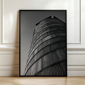 May include: A black and white framed wall art print featuring a low-angle perspective of a cylindrical metal grain silo. The structure displays vertical corrugated panels and a curved silhouette against a plain, solid gray sky background.