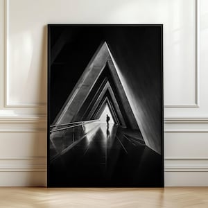 Könnte beinhalten: Ein schwarz-weiß gerahmter Wandkunstdruck mit einer perspektivischen Ansicht eines dreieckigen architektonischen Tunnels. Eine kleine Silhouette steht im hellen Licht am Ende des Korridors, der glatte, dunkle Böden und Wände aufweist.