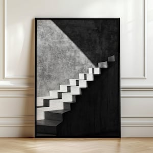 Puede incluir: Una impresión artística enmarcada en blanco y negro que muestra una escalera ascendiendo en diagonal. La imagen presenta un fuerte contraste entre luces y sombras, con los escalones parcialmente iluminados sobre un muro de hormigón texturizado.