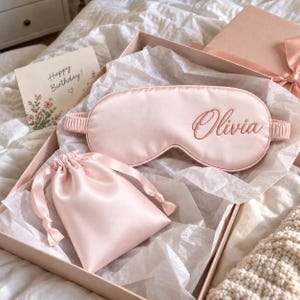 May include: A light pink sleep mask with the name Olivia embroidered in a darker pink script font, accompanied by a matching light pink drawstring pouch, both presented inside a gift box with a Happy Birthday card.