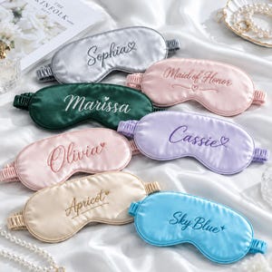 May include: A collection of seven satin sleep masks in various colours including silver, pink, dark green, light pink, lavender, beige, and blue. Each mask features embroidered names or text such as Sophia, Maid of Honor, Marissa, Cassie, Olivia, Apricot, and Sky Blue.