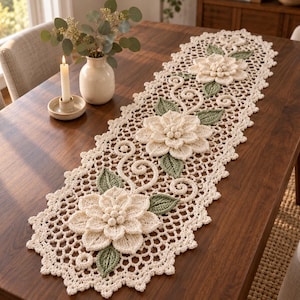 May include: A cream-colored crocheted table runner featuring three large, dimensional floral motifs with layered petals and small central spheres. The runner includes sage green crocheted leaves and intricate scrollwork, finished with a scalloped edge.