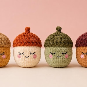 Può includere: Quattro peluche all'uncinetto a forma di ghianda disposti in fila. Ogni giocattolo presenta un cappuccio testurizzato, un corpo liscio, occhi chiusi, un piccolo sorriso e due guance circolari rosa. I colori includono beige, crema, verde salvia e marrone con cappucci rosso scuro.