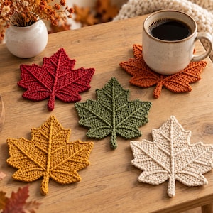 May include: A set of five crochet coasters shaped like maple leaves in red, orange, green, yellow, and cream colors. One orange coaster supports a ceramic mug filled with black coffee. The coasters feature detailed stitched veins and textures.