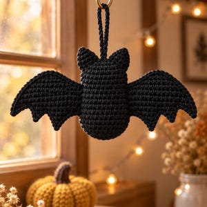 Pode incluir: Um ornamento de morcego em crochê preto com uma superfície de fio texturizado, orelhas pontiagudas e asas largas e recortadas. O morcego está suspenso por uma alça trançada preta a condizer, concebida para ser pendurada.