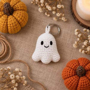 May include: A white crocheted ghost keychain featuring two small black embroidered eyes and a simple black stitched smile. The ghost is crafted with a soft yarn texture and includes a silver-toned metal split ring attached to the top for hanging.