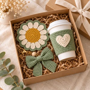 Può includere: Una scatola regalo contenente un sottobicchiere all'uncinetto a forma di margherita con petali gialli e bianchi, una fascia per tazza all'uncinetto verde salvia con un cuore bianco e un portachiavi a fiocco all'uncinetto verde salvia.