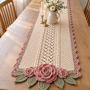 Puede incluir: Un camino de mesa rectangular de ganchillo color crema con un patrón calado central, bordeado por un ribete verde salvia y un borde festoneado rosa empolvado. El camino está decorado con rosas de ganchillo tridimensionales y hojas verdes.