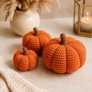 May include: Three crocheted pumpkins in varying sizes, each featuring a vibrant orange body with a textured, ribbed surface and a small, dark brown crocheted stem at the top. The pumpkins are arranged together, showcasing their rounded, soft forms.
