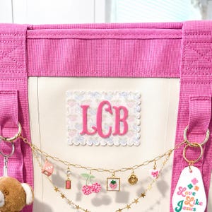 Op de afbeelding: Een felroze tas met een crèmekleurig middenpaneel voorzien van een geschulpte patch met de roze monogramletters LCB. Een goudkleurige ketting hangt aan de voorzijde, versierd met bedels waaronder kersen, een aardbei en een label met de tekst Love Like Jesus.