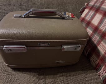 May include: A vintage taupe-colored Samsonite train case with a chrome handle and latches. The case has a rectangular shape and a textured surface. A red tag hangs from the handle. The case is on a brown surface.
