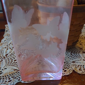 May include: A pink glass vase with a square shape and etched butterfly designs. The vase has a slightly flared opening and is sitting on a table with a lace doily.