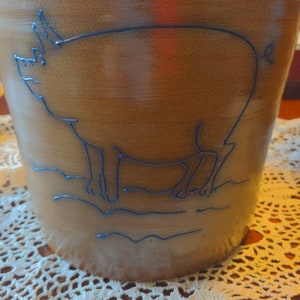 May include: A brown ceramic pitcher with a black outline of a pig on the front. The pitcher is sitting on a white crocheted doily.
