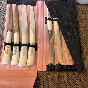 May include: A set of six knives in a pink leather case with a black crocodile-textured flap. The knives have white handles with gold floral designs and silver blades. The knives are wrapped in paper.