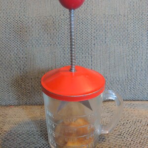 May include: A red and clear glass apple corer with a spring-loaded plunger and a red knob on top. The corer has a handle and a measuring cup on the side with markings for 1/4 cup, 1/2 cup, 3/4 cup, and 1 cup.