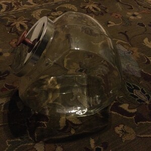 May include: A clear glass jar with a silver lid and a brown knob. The jar is sitting on a patterned rug.