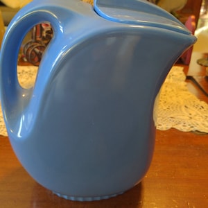 May include: A vintage, powder blue ceramic pitcher with a curved handle and a matching lid. The pitcher has a rounded body and a scalloped base. The design is streamlined and classic, suitable for serving beverages.