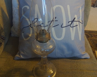 May include: A clear glass oil lamp with a decorative glass chimney and a metal burner. The lamp is sitting on a gray couch with a pillow that says "Snow Let it" in white lettering.