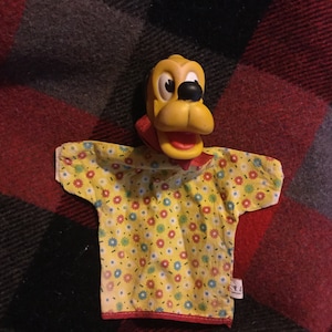 May include: Yellow and black puppet with a floral print shirt. The puppet is a cartoon dog with a red mouth and black eyes.