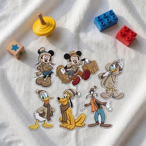 Könnte beinhalten: Eine Sammlung von sechs gestickten Aufnähern mit Disney-Figuren wie Mickey Maus, Minnie Maus, Donald Duck, Daisy Duck, Pluto und Goofy, alle in Winterkleidung, begleitet von einem gelben Kreisel und zwei Kunststoff-Bausteinen.