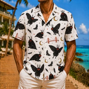May include: A short-sleeved button-down shirt in off-white featuring an all-over print of black moths with red eyes, golden crescent moons, small stars, feathers, and red illustrations of the Golden Gate Bridge across the fabric.