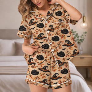 Op de afbeelding: Een beige pyjamaset bestaande uit een shirt met korte mouwen en knoopsluiting en een bijpassende short. De stof heeft een herhalend patroon van zwarte katten, heksenhoeden met maansikkels, kleine vleermuizen en herfstkleurige bladeren.