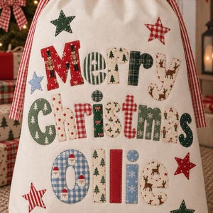 May include: A cream-coloured fabric drawstring gift bag featuring the appliqued text Merry Christmas Ollie. The letters are decorated with various patterns including red nutcrackers, green trees, reindeer, and blue Santa Claus motifs.
