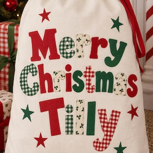 May include: A cream-coloured fabric drawstring gift sack featuring the text Merry Christmas Tilly in a mix of red, green, and patterned fabric letters. The bag is decorated with red and green stars and includes a red ribbon drawstring closure.