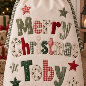 May include: A cream-coloured fabric drawstring gift bag featuring the text Merry Christmas Toby in multi-coloured, patterned fabric letters. The bag is decorated with various star-shaped fabric appliques and has green and white checkered drawstrings.