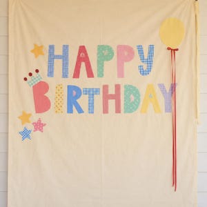 May include: A cream-coloured fabric banner featuring the text HAPPY BIRTHDAY in multicoloured, patterned fabric letters. The design includes a yellow fabric balloon with red ribbons, a small crown, and several stars in blue, yellow, and pink.