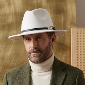 Può includere: Un cappello fedora bianco con una fascia nera, un maglione a collo alto color crema e un blazer verde oliva. Il blazer presenta un tessuto testurizzato, revers classici e due bottoni frontali. L'insieme è indossato con jeans blu scuro.