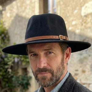 May include: A black fedora hat with a tan leather band and gold-tone buckle, a charcoal grey herringbone blazer, a light grey button-down shirt, and a wristwatch with a tan leather strap and a round face with a dark dial and gold-tone hands.