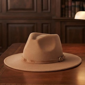 Puede incluir: Un sombrero fedora de color tostado con ala ancha y una banda tonal a juego que presenta un pequeño detalle de hebilla metálica. El sombrero tiene una copa estructurada con un pliegue central y está hecho de un material liso tipo fieltro.