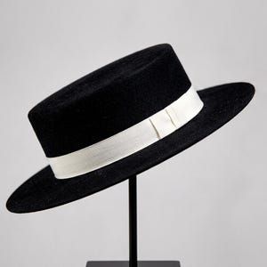 Può includere: Un cappello nero a tesa piatta con una falda larga e rigida e un nastro bianco a contrasto avvolto attorno alla base della corona. Il cappello presenta una corona cilindrica strutturata e una parte superiore piatta e uniforme.