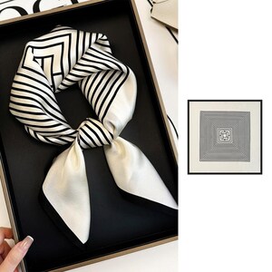 Peut inclure: Un foulard carré en soie arborant un motif géométrique noir et blanc composé de carrés concentriques et de lignes parallèles. Le foulard est présenté noué et à plat, révélant un design répétitif en forme de labyrinthe avec un bord noir uni.
