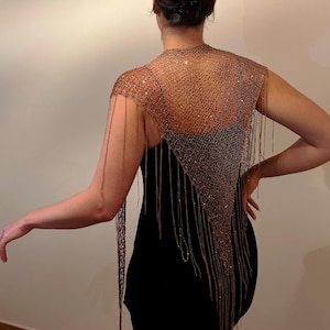 May include: A metallic copper-colored mesh shawl featuring a fine, open-knit texture and long, dangling fringe strands that drape over the shoulders and back. The garment is worn over a simple black sleeveless dress, creating a layered appearance.