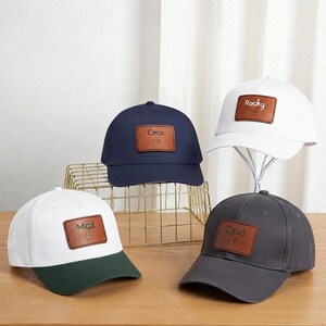 Può includere: Quattro cappellini da baseball con toppe rettangolari in pelle marrone con nomi ricamati e impronte di zampe. Un cappellino è blu navy, uno bianco, uno grigio scuro e uno bianco con visiera verde scuro. Sono visibili i nomi Max, Coco, Rocky e Zeus.