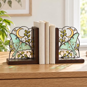 May include: A pair of L-shaped bookends featuring dark brown wooden bases and decorative metal panels. Each panel displays a stained glass-style luna moth in pale green, a yellow crescent moon, and green leafy vines with small gold accents.