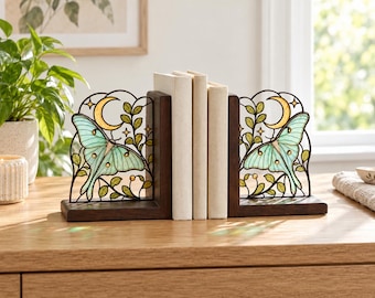 May include: A pair of L-shaped bookends featuring dark brown wooden bases and decorative metal panels. Each panel displays a stained glass-style luna moth in pale green, a yellow crescent moon, and green leafy vines with small gold accents.