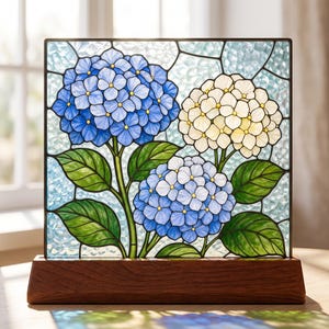 Puede incluir: Un panel cuadrado de vidriera con tres racimos de flores de hortensia en tonos azul, azul claro y crema, sobre un fondo azul claro texturizado con hojas verdes, montado sobre una base rectangular de madera de color marrón oscuro.