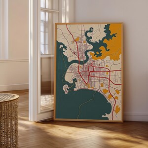 May include: A rectangular framed wall art print featuring a stylised city map. The map displays a network of thin black roads and prominent red arterial routes set against a cream background, with deep teal water and mustard yellow land areas.