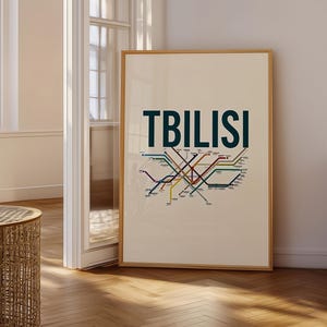 May include: A vertical rectangular framed print featuring the word TBILISI in large dark blue capital letters above a stylised multi-coloured transit map with various intersecting lines and small text labels on a light cream background.