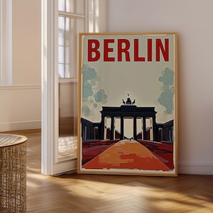 May include: A vertical rectangular poster in a light wood frame featuring a stylised illustration of the Brandenburg Gate in Berlin. The word BERLIN is printed in large red capital letters at the top, with a dark gate and orange path below.